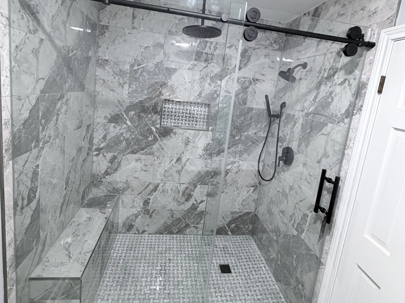 Custom tile shower with marble-look walls, built-in bench, basketweave floor and black sliding glass door